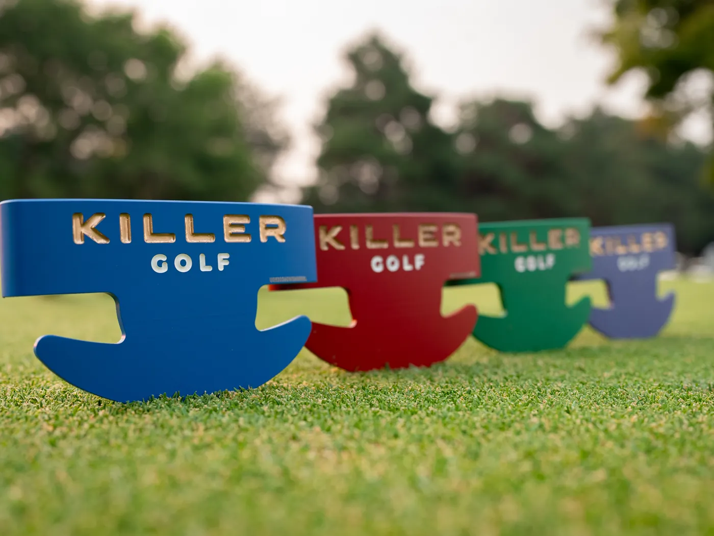 Four Killer Golf putter heads in blue, red, green and lavender, each with the KILLER GOLF logo, standing in a row on a golf green.